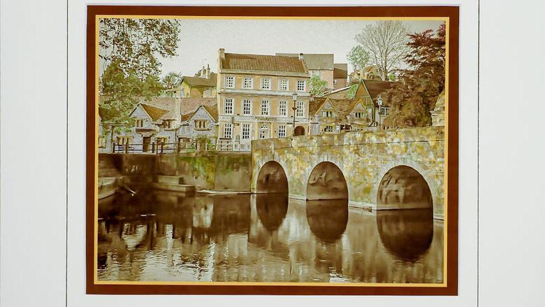 Hand-colored, monochrome photograph of stone bridge and building by Frank Santimauro